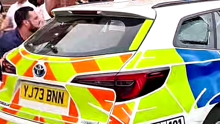 A man standing by the rear left window of a police car, which has its window on the other side smashed. His body is facing to his left.