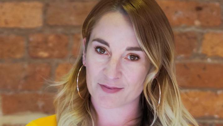 A professional headshot of a woman with long blonde hair standing in front of a brick wall.