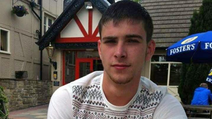 A photo of James sitting in a beer garden outside a pub. He has short dark hair and is clean-shaven. He is looking directly at the camera, wearing a white jumper with a pattern on the front but not the sleeves.