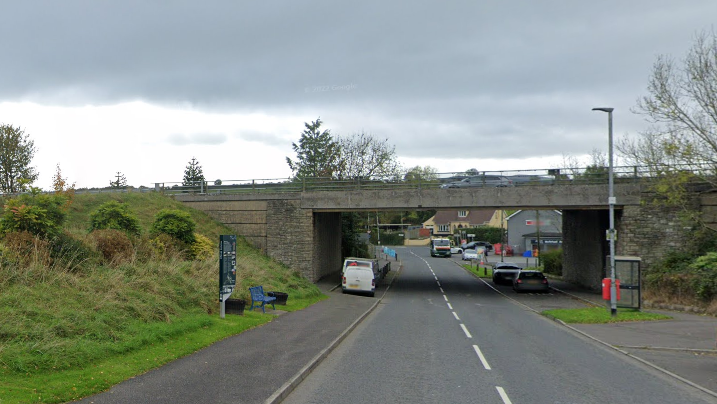 A Google Maps screen grab of Fyfin Road near Victoria Bridge.  It shows a single carriageway road running under a concrete road bridge.  There are parked vehicles, a phone box, and a metal bench in the vicinity. 