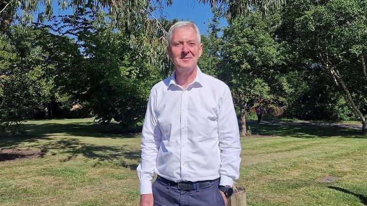 Tim Oliver looking at the camera. He is wearing a grey shirt and blue trousers. Grass and trees in the background.