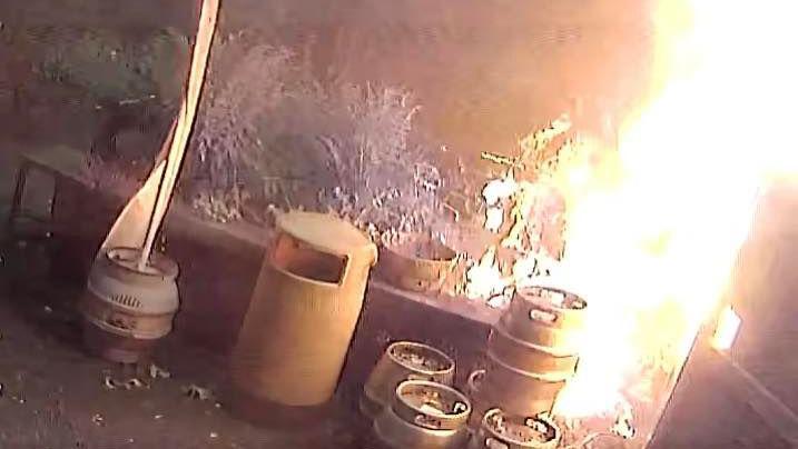 CCTV footage of the fire. There is a bin and multiple kegs stacked on top of each other. The fire is on the right side of the image and goes up to the top of the picture.