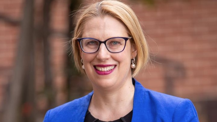 A blonde woman wearing a black shirt, blue blazer and eye glasses looks at the camera. She is wearing pearl earrings and lipstick