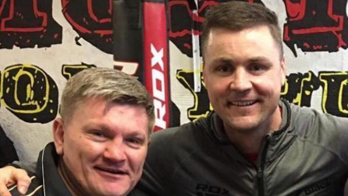 Ricky Hatton and Thomas McDonagh smile as they pose together in a gym with a branded wall behind them. They are wearing dark casual sporting tops.