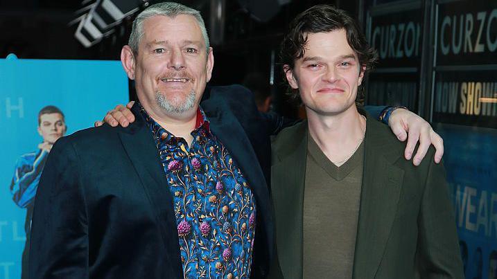 John Davidson, in a black jacket and thistle-patterned shirt puts his arm around actor Robert Aramayo who wears a dark green jacket and olive sweater. Both smile for the camera.