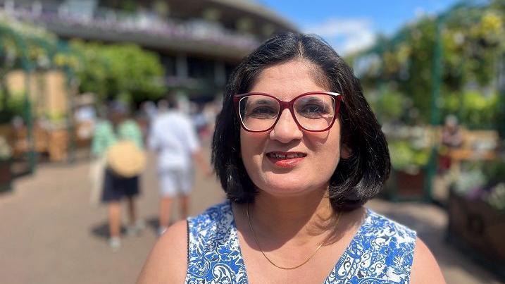 Kam Soomal is wearing a blue dress and a necklace. She has short black hair and glasses. There are people walking around in the background.