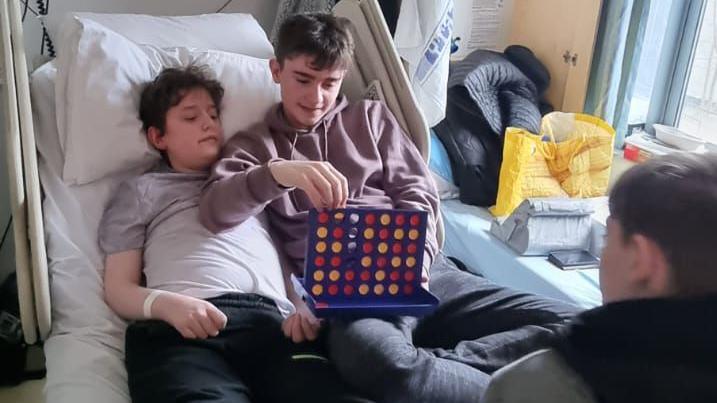 Kyle with his brother Liam lying in a hospital bed. Liam is putting the counters in a Connect Four grid as Kyle decides where they should go. The opponent is sitting facing away from the camera. 