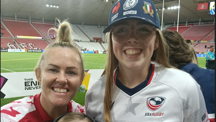 A rugby player in a red top with her blonde hair scraped up on top of her head. She is standing next to a taller teenage girl with long blonde hair, glasses, a blue baseball cap and white ruby shirt with USA rugby written on the badge of an eagle, rugby ball and red stripes. They are standing beside the pitch in a stadium with red seats. Both are smiling.