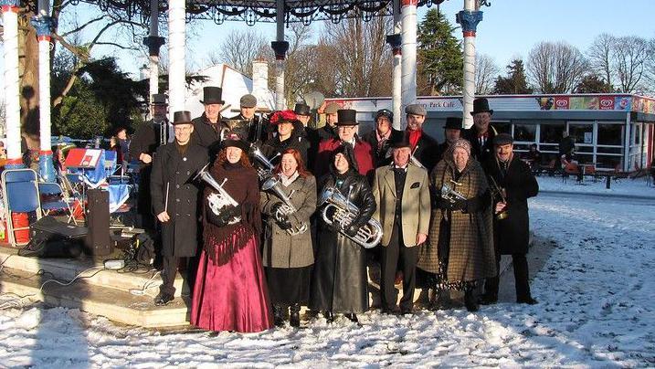 A group of people with instruments including silver trombones are stood next to a bandstand in a park. There is snow on the ground and the group of people are wearing top hats and old fashioned style clothing. One lady in the front of the image wears a long red dress with a shawl. 