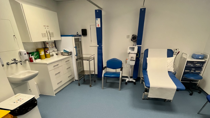 An image of a clinical room in Honiton Surgery. It is clean with new storage units, equipment and a sink. There is a blue bed on the right with a blue chair to the left of it. 