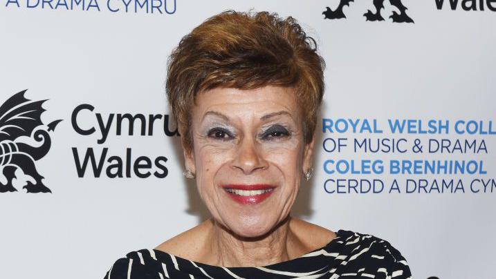 Iris Williams smiling at the camera. She wears a black and white top. She is standing in front of a white background with Cymru Wales, in black writing, and Royal Welsh College of Music & Drama, in blue writing, behind her.
