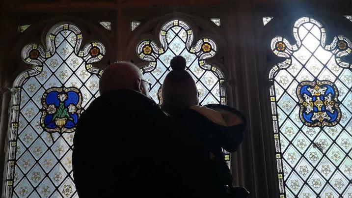 Mr Berry and one of his grandchildren pictured from behind, looking up at a stained glass window in a church. They are in silhouette and there is bright light shining through the three ornate panels of glass. 