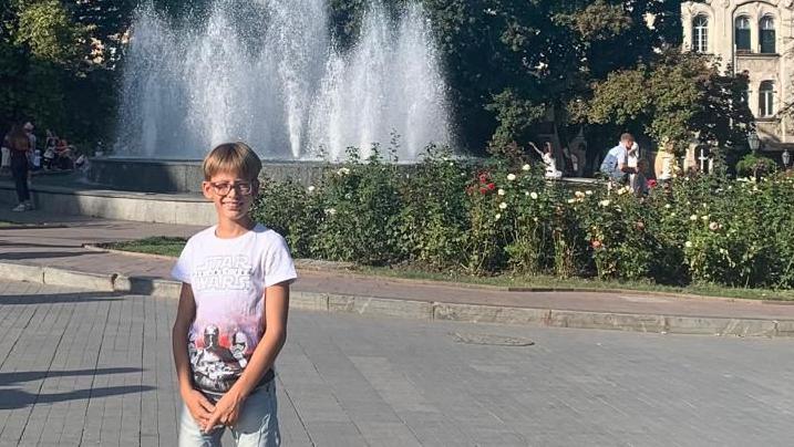 A boy with blonde hair and wearing a Star Wars T-shirt and jeans stands in front of a fountain in a city square, with trees and flowers in the background.