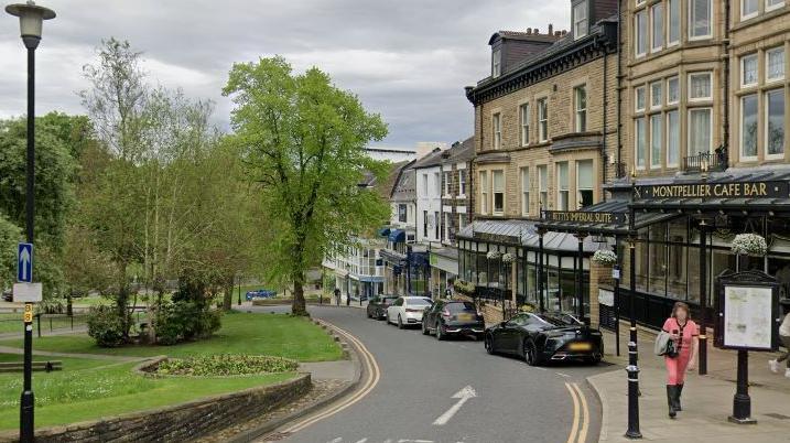 View down Montpellier Parade in Harrogate - a row of shops and cafes with flats above them to the right of the picture with a grassed area and trees on the other side of the road