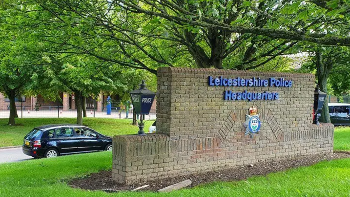 Leicestershire Police headquarters - with a sign showing the force's name over a crest on a brick wall.  Blue police-style lanterns sit on either side of the sign.  There is a black car and trees in the background.