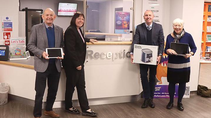 Two men and two women, all wearing smart clothes, stand in a modern-looking reception area of a library. They are holding electrical appliances and smiling for the camera