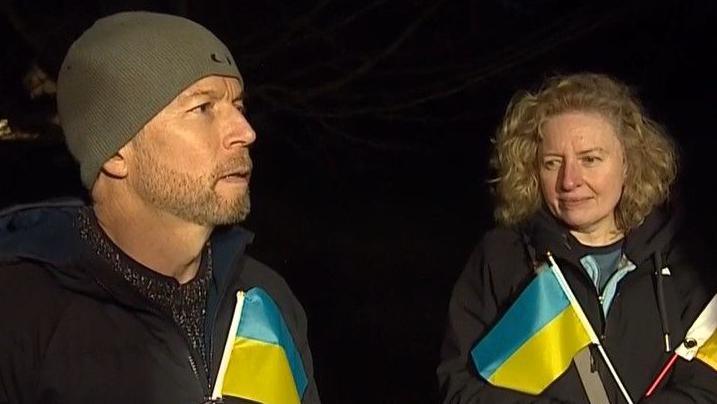 Man with woollen hat and woman with blond wavy hair hold Ukrainian flags. 