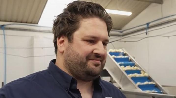 Dom Horabin standing in a potato factory. There is a conveyor belt with potatoes in the background. Dom is wearing a navy shirt.