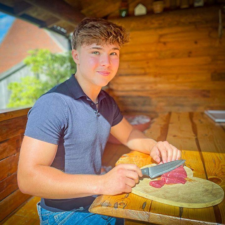 Nat Walney, who has brown hair and blue eyes, sat in a wooden cabin, sat on a wooden bench and eating raw steak from a plate on a wooden table. He is wearing a navy T-shirt and looking at the camera slightly smiling.