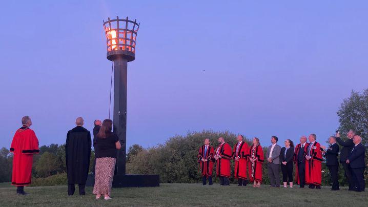 A group of people standing around a beacon being lit in a field at dusk.