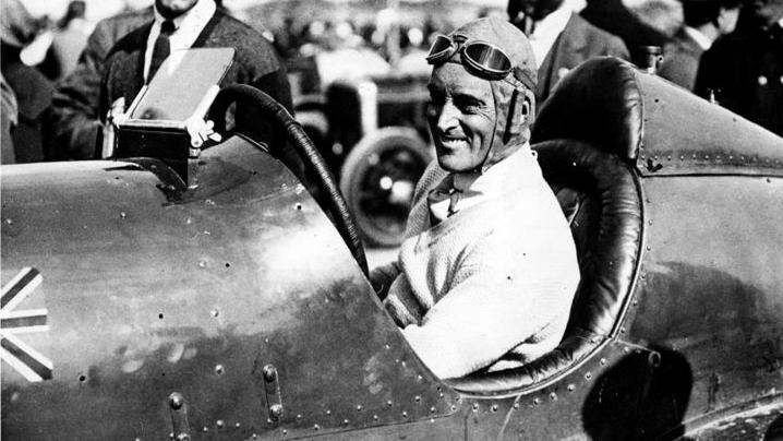In black and white, Malcolm is behind the wheel of Blue Bird. He is wearing a light coloured v-neck jumper , a leather aviator helmet with flaps over his ears and goggles on his forehead. Clearly visible are the rudimentary rivets used to hold together Bluebird's bodywork.