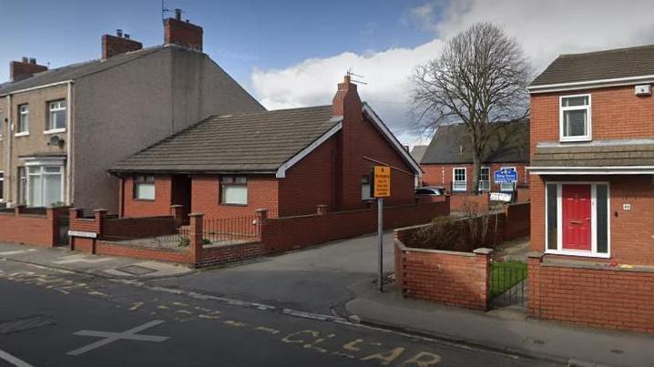 A bungalow next to the entrance to a school. You can just see the blue sign to the school behind the bungalow.