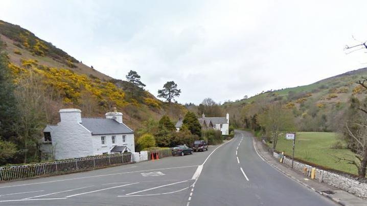 The AS at the junction with Ballig Bridge. There is a white cottage on the left and green fields on the right and there are hills on both sides.