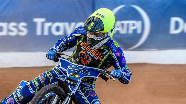 A speedway racer steers his bike around a corner on a mud track. He wears a bright green helmet and blue racing suit with various sponsors written on it.