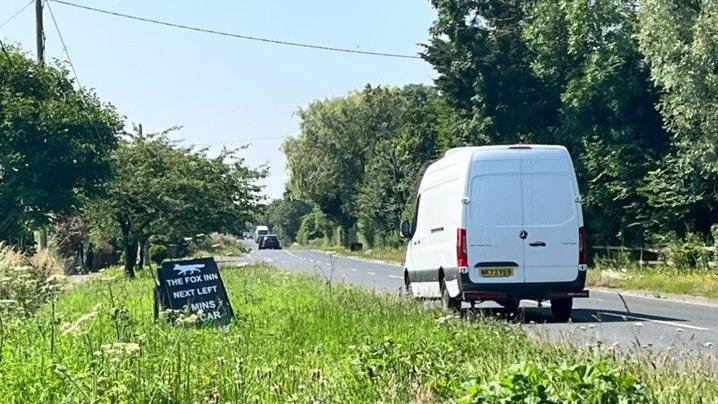 Section of A12 road showing pub sign on grassy verge and white van on carriageway