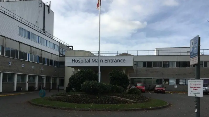 The main entrance of the Princess Elizabeth Hospital main entrance. There is a landscaped mini roundabout with a flagpole at the centre in front of the entrance. The building is low and grey. A few cars are parked in spaces immediately outside the building. 