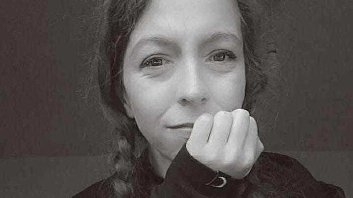 A black and white selfie of a woman with long hair in two plaits. She has her chin resting in her hand and is smiling.