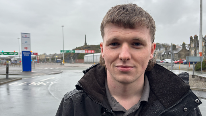 A man is stood front of a port. It is a wet day. He is looking at the camera and has dark hair in the shape of a mullet. He is wearing a black jacket over a grey shirt.