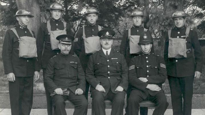 A group of men in wartime police uniform, some sitting and others standing.
