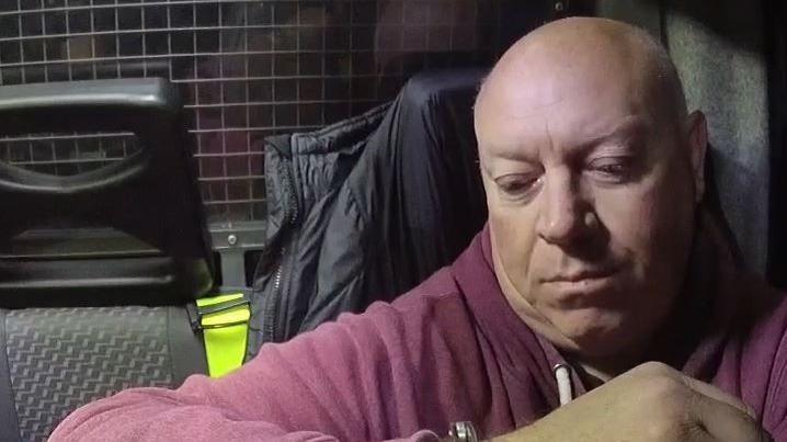 A police image of Andrew Griggs who is sitting in the back of a car. He is looking at his arm, on which a handcuff is just visible and is wearing a dark red hoodie.
