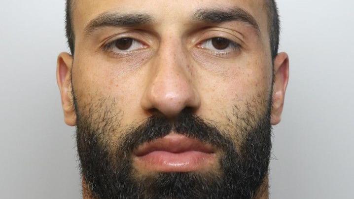 A mugshot of a young man with short hair and a dark beard. He is staring directly at the camera.