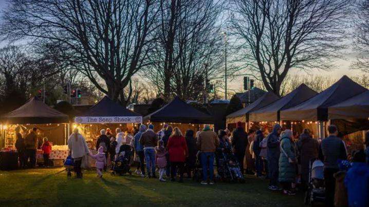 Crowds of people look at Christmas market stalls on a grassy lawn at twilight.