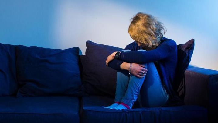 Blonde woman sits in a dark room, cradling her legs with her arms.