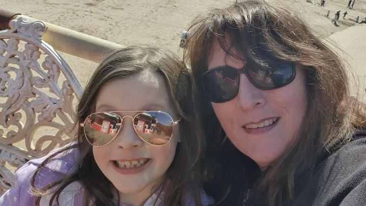 Sandra with her daughter Beatrice. They are taking a selfie by the seaside. There is a beach behind them. They are both wearing sunglasses. 