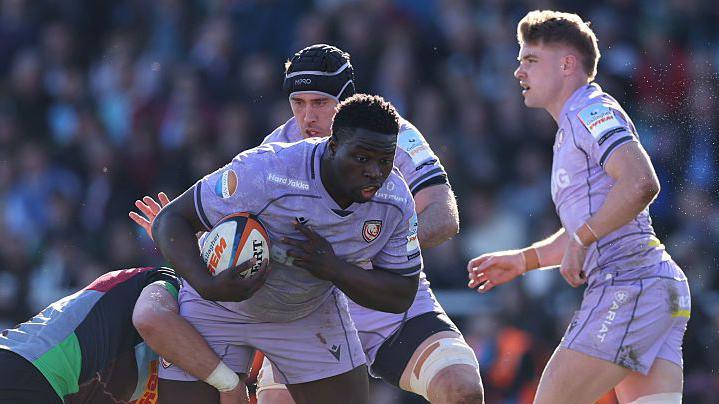 Afo Fasogbon holds the ball under his right arm a Harlequins player wraps around his leg in an attempt to make a tackle