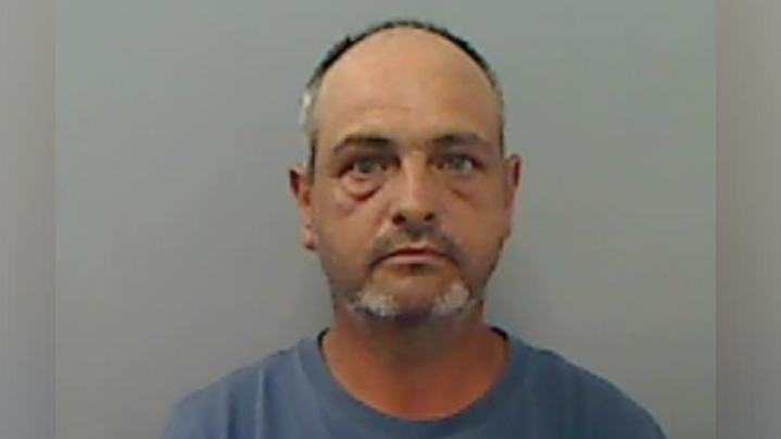 Anthony Allen, with a balding head, and grey moustache and beard, in a police mugshot photo. He is wearing a blue Adidas top.