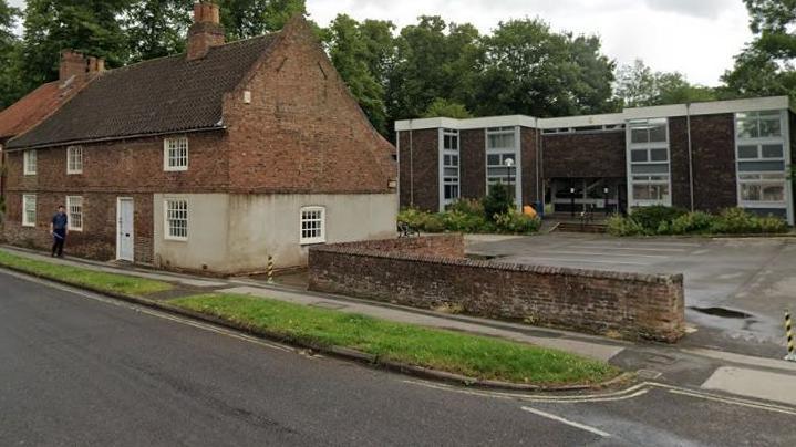 Two contrasting buildings face an empty asphalt road: In the foreground on the left behind a strip of grass is an 18th Century steep roofed building with small windows. To the right of it is a car park, at the rear of which is a 1960s brick office building.