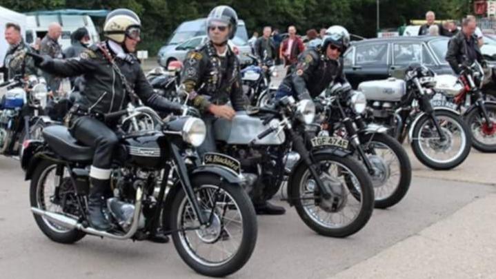 LIne of old-style motorbikes in a car park, all black in colour with large headlamps. The bikers are wearing leathers and helmets and are either sitting on their bikes or standing beside them. A car is visible to the right and a white van to the left.