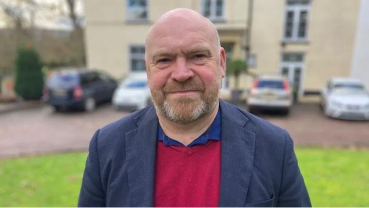 Somerset Council Leader Bill Revans stood outside a cream coloured building. He is wearing a blue suit jacket and a red jumper. 