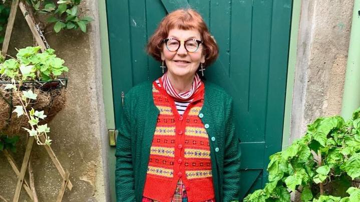 A woman with short red hair and black glasses stands in front of a green door. She is wearing a green cardigan with a knitted red and yellow vest underneath. A flower hanging basket sits to her left with a bush to her right. 