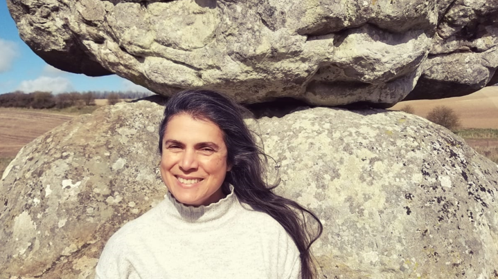 A close-up of Deborah in a field standing in front of a two large stones balancing on top of one another. She is smiling and her long, dark hair appears to be blowing in a gentle wind.