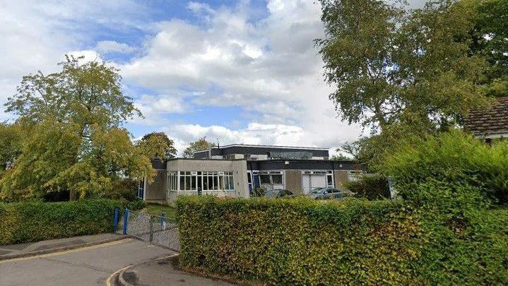 A concrete school building behind a leafy hedge and gate