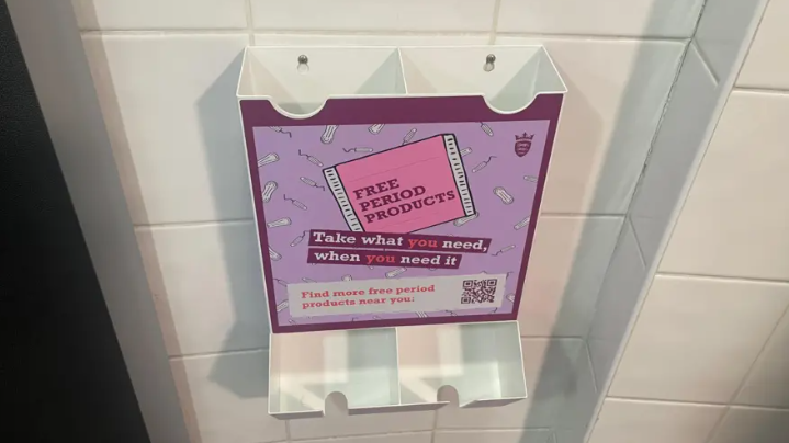 A plastic dispenser of free period products. The dispenser has a large sticker on the front that says 'free period products take what you need, when you need it'