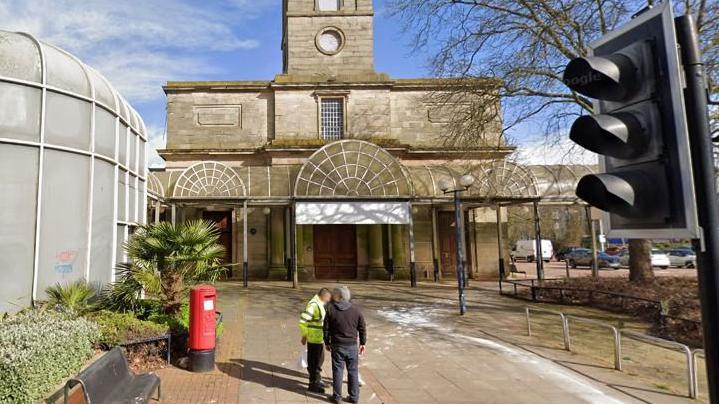 A large white-stone building with two people standing on the pavement in front of it.