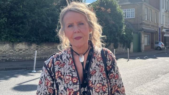 Jilly Fernandez, standing outside, by the side of a road, with bollards behind her, and shops in the distance. She has long blonde hair, tied back, is wearing a large silver necklace and a flower patterned jacket.