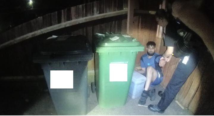 A police officer holding the arm of Mohammed Mehtab, who is on the floor behind a green wheelie bin